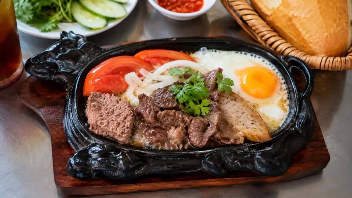 Bò Né vietnamesisk fräsande nötkött i gjutjärnspanna med stekt ägg, paté, lök och tomat serverat med baguette